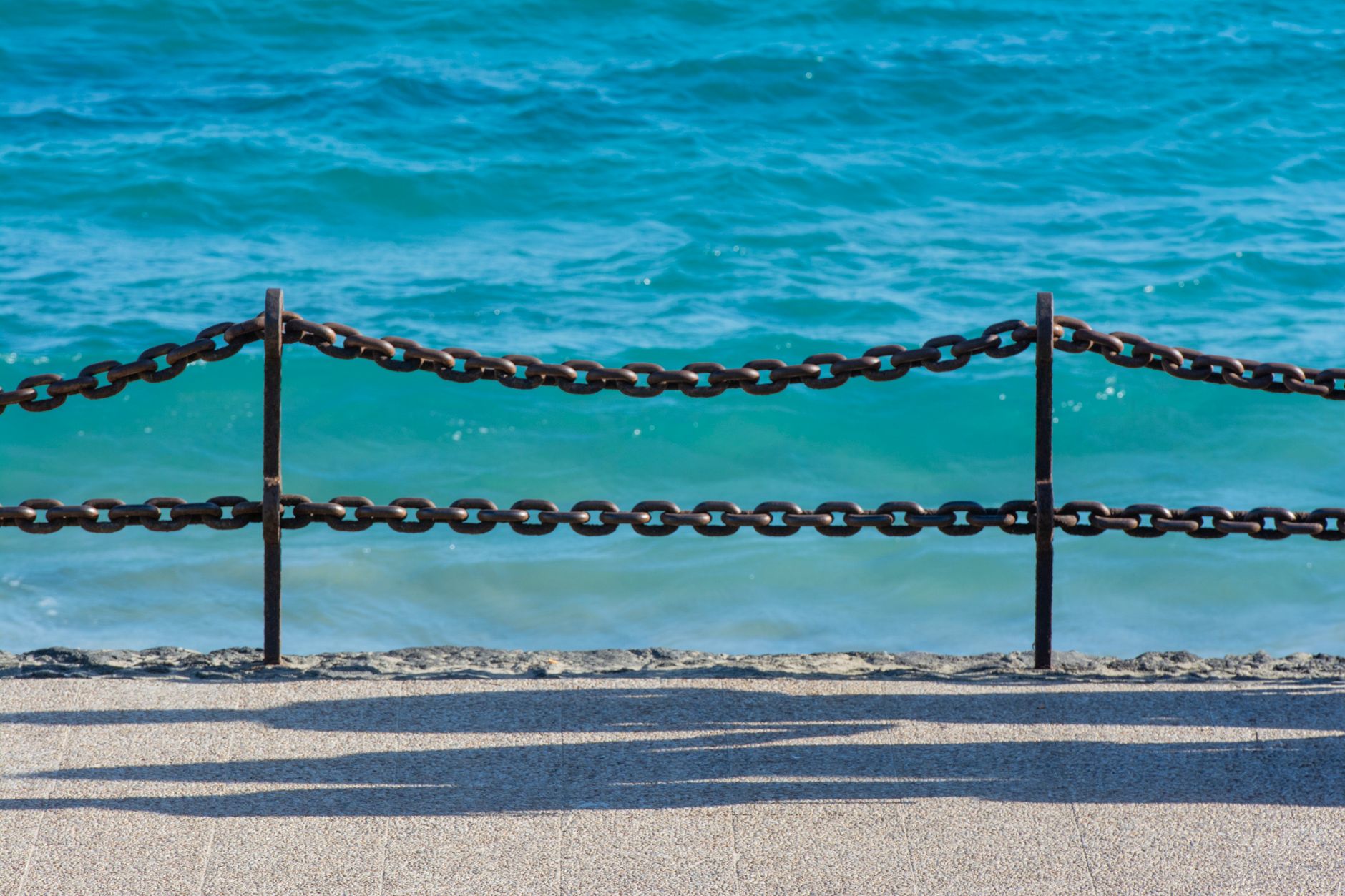 Rusty chain fence lining a sunny seaside sidewalk with clear turquoise ocean in the background.
