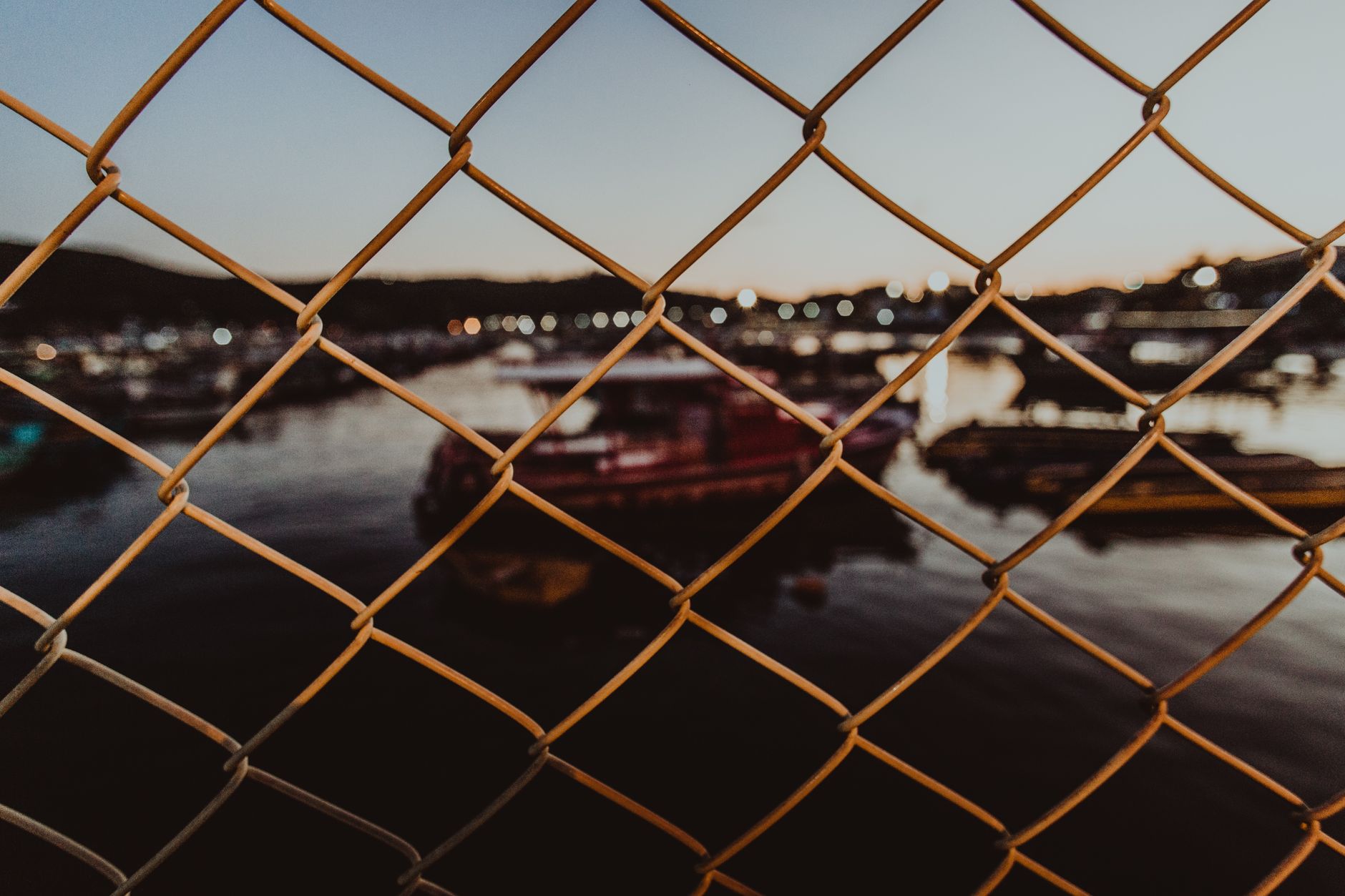 Through chain link fence of various ships floating on sea water near harbor against cloudless evening sky in illuminated coastal town